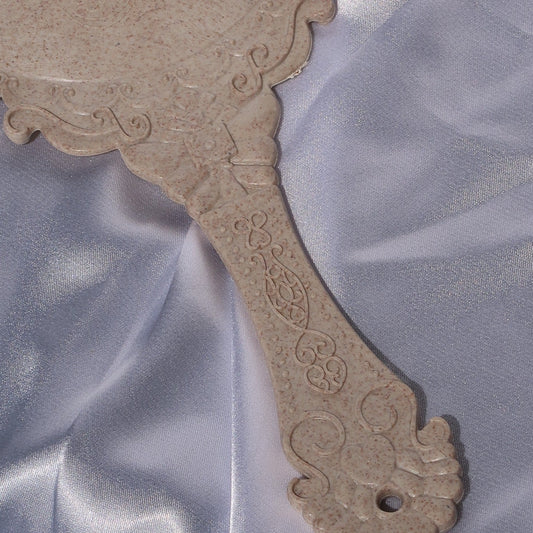 Close-up of the ornate floral detailing on the back and handle of a beige vintage-style mirror.