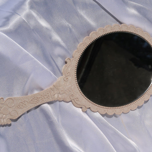 Front view of a beige vintage-style handheld mirror with an ornate frame and round mirror.